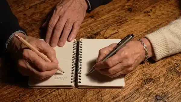 Two pocket notebooks side by side, a man's hand and a woman's hand each writing one line