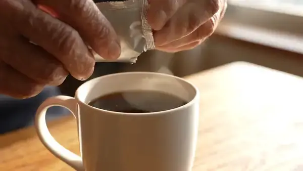 Torn foil sachet squeezed slowly into a mug of black coffee, the pale paste swirling down